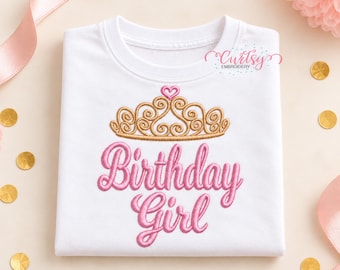 May include: A white short-sleeved t-shirt featuring an embroidered gold tiara with a small pink heart at the top. Below the tiara, the text "Birthday Girl" is embroidered in pink script lettering. The shirt includes a small logo.