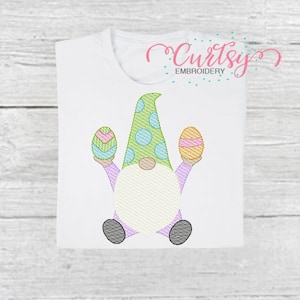 K&ouml;nnte beinhalten: Ein wei&szlig;es T-Shirt mit einem gestickten Design eines Zwergs, der zwei Ostereier h&auml;lt. Der Zwerg tr&auml;gt einen gr&uuml;n-blauen Hut mit wei&szlig;en Punkten. Die Eier sind mit rosa, gelben und gr&uuml;nen Streifen verziert.