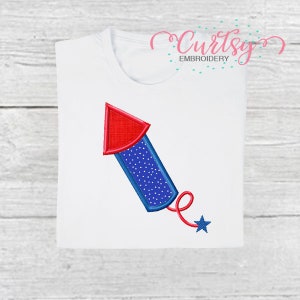 May include: White t-shirt featuring a patriotic embroidered design of a red and blue firework with a star tail. The firework has a red tip, a blue body with white dots, and a blue outline. The shirt has the text "Curtsy Embroidery".