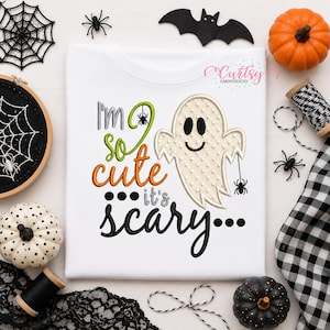 May include: A white t-shirt featuring an embroidered design of a cream-coloured ghost with a textured surface. The text reads I'm so cute it's scary, with a small spider hanging from the ghost and another spider integrated into the letter O.