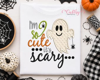 May include: A white t-shirt featuring an embroidered ghost and the text "I'm so cute it's scary". The ghost is cream-colored with a textured surface, holding a small black spider. The lettering uses green, orange, and black thread.