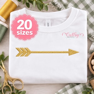 Może przedstawiać: Biały t-shirt ze złotym haftowanym wzorem strzałki na środku. Różowa, karbowana plakietka w lewym górnym rogu zawiera napis 20 sizes. Logo Curtsy Embroidery jest nadrukowane na różowo w prawej górnej części koszulki.