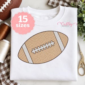 May include: A white t-shirt featuring a central machine embroidery design of a brown American football with white laces and stripes. A pink scalloped badge on the upper left displays the text 15 sizes, and the Curtsy Embroidery logo is visible.