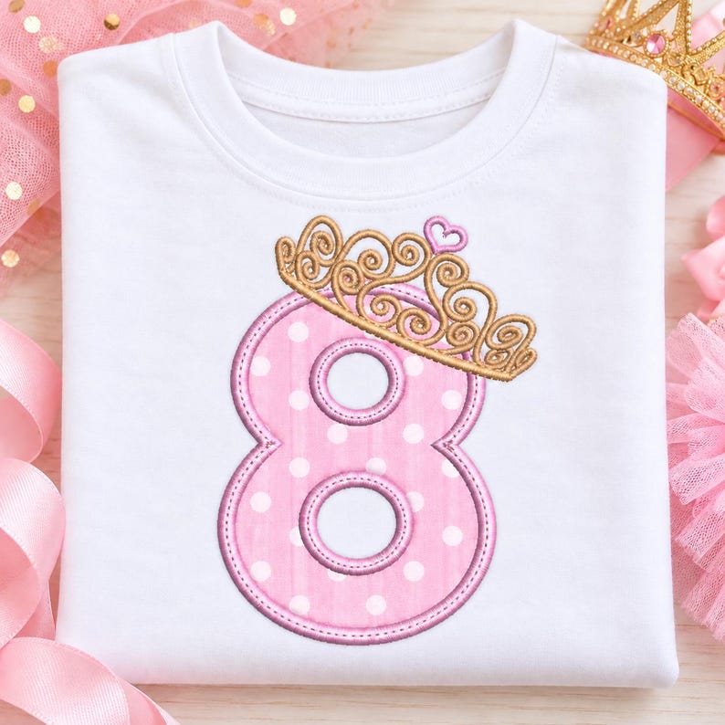 May include: A white t-shirt featuring an embroidered pink number eight with white polka dots. The number is topped with a gold-colored crown design that includes a small pink heart detail at the top center of the crown embroidery.