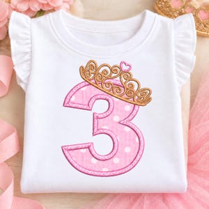 May include: A white short-sleeved shirt with ruffled sleeves featuring an embroidered pink number three with white polka dots. A gold-colored crown with a small pink heart sits atop the number three, showcasing detailed stitching.