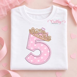 May include: A white t-shirt featuring an embroidered pink number five with white polka dots. The number is topped with a gold-colored crown and a small pink heart. The text Curtsy Embroidery is printed in the upper right corner of the shirt.