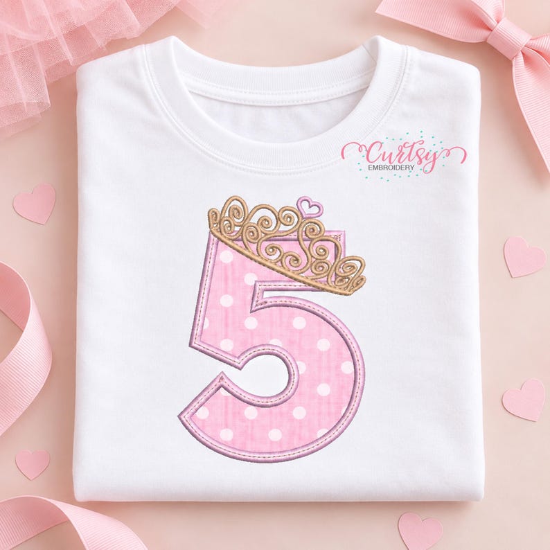 May include: A white t-shirt featuring an embroidered pink number five with white polka dots. The number is topped with a gold-colored crown and a small pink heart. The text Curtsy Embroidery is printed in the upper right corner of the shirt.