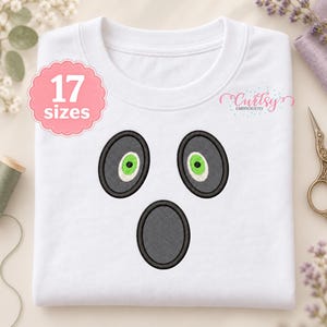 Könnte beinhalten: Ein weißes T-Shirt mit einem gestickten Geistergesicht. Das Design besteht aus zwei dunkelgrauen ovalen Augen mit hellgrünen Iris und einem dunkelgrauen ovalen Mund. Ein rosa gewelltes Abzeichen zeigt 17 sizes und der Text Curtsy Embroidery ist sichtbar.