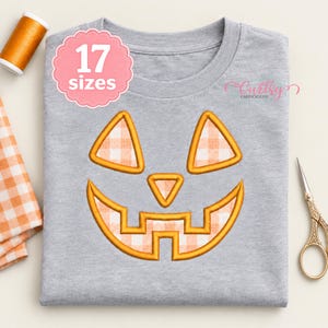 May include: A heather gray t-shirt featuring an embroidered jack-o'-lantern face with orange and white gingham fabric fill. The design includes two triangular eyes, a small triangular nose, and a wide, jagged mouth. A pink badge reads 17 sizes.