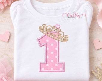 May include: A white short-sleeved shirt featuring an embroidered pink number one with white polka dots, topped with a gold crown and a small pink heart. The text Curtsy Embroidery is printed in the upper right corner of the shirt.