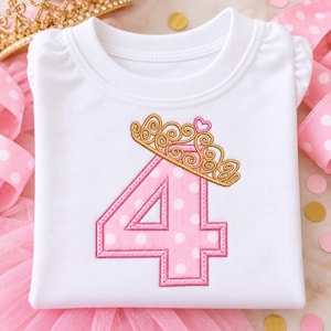 May include: A white short-sleeved shirt featuring a large pink number four with white polka dots. The number is topped with an embroidered gold crown design that includes a small pink heart at the center. The shirt has gathered shoulder details.