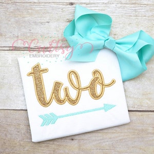 May include: A white onesie with the word "two" in gold glitter lettering and a turquoise arrow underneath. A turquoise bow is on top of the onesie.