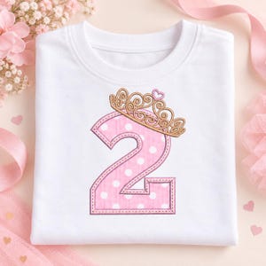 May include: A white t-shirt featuring an embroidered number two in pink with white polka dots. The number is topped with a gold-colored embroidered crown design that includes a small pink heart detail at the very top center.