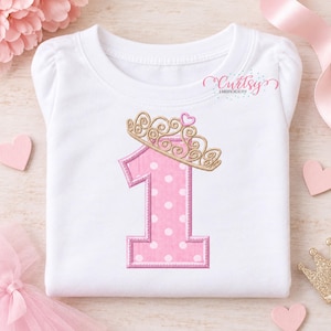 May include: A white short-sleeved shirt featuring an embroidered pink number one with white polka dots, topped with a gold crown and a small pink heart. The text Curtsy Embroidery is printed in the upper right corner of the shirt.