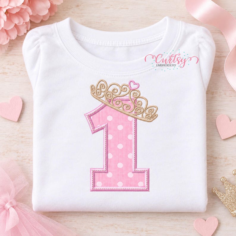 May include: A white short-sleeved shirt featuring an embroidered pink number one with white polka dots, topped with a gold crown and a small pink heart. The text Curtsy Embroidery is printed in the upper right corner of the shirt.