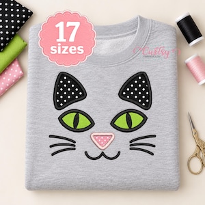 Pode incluir: Uma camisola cinzenta mesclada com uma cara de gato bordada com olhos verde-brilhantes, orelhas com bolinhas pretas e brancas e um nariz com bolinhas cor-de-rosa. O texto 17 sizes está num círculo rosa recortado sobre o gato.