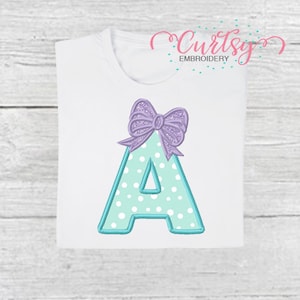 May include: A light blue letter A with white polka dots and a light purple bow on top. The letter is embroidered on a white t-shirt.