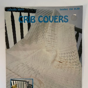 May include: A vintage Leisure Arts leaflet titled "Crib Covers" with a white knitted blanket draped over a wooden chair. The leaflet features a photo of a crib with two stuffed animals and the text "8 To Knit And Crochet".