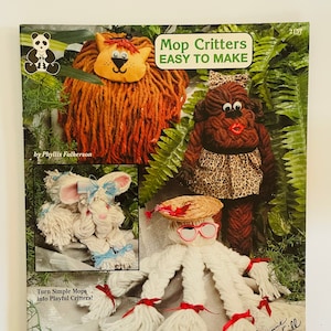 Puede incluir: Una portada de libro con el título "Mop Critters Easy To Make" y una imagen de cuatro animales diferentes hechos con mopas. Los animales son un león, un mono, un conejo y una criatura con un sombrero de paja y gafas. La portada del libro también incluye el texto "Just Follow Me... Never-Fail Patterns and Instructions" y "No. 2157".
