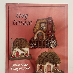 May include: A book titled "Cozy Cottage" with illustrations of three small, wooden houses with moss roofs. The book is by Susan Roach and Emily Dinsdale.