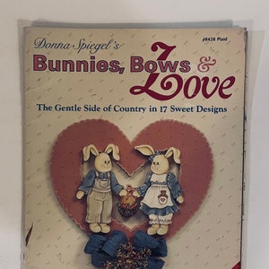 May include: A book cover with a pink heart and two bunnies holding a basket of flowers. The title is "Bunnies, Bows & Love" and the subtitle is "The Gentle Side of Country in 17 Sweet Designs". The book is by Donna Spiegel and is a Plaid publication.