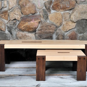 May include: A set of two rectangular tables featuring light-colored wooden tops with a dark rectangular inlay in the center. The tables have thick, dark brown wooden legs joined to the top with visible dark circular fasteners at each corner.