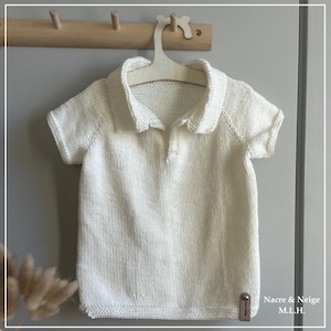May include: White knitted baby polo shirt with a button closure and short sleeves. The shirt is hanging on a wooden hanger. The tag on the shirt reads "Nacre & Neige M.L.H."