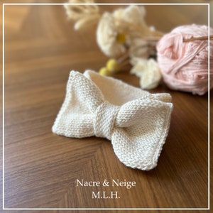 May include: A white knitted bow tie with a textured surface. The bow tie is on a wooden surface. Nacre & Neige M.L.H.