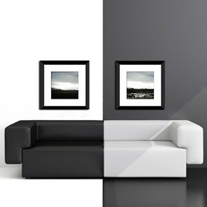 May include: A black leather sofa with a white cushion sits in front of a white wall and a black wall. Two black framed photographs hang on the walls above the sofa.