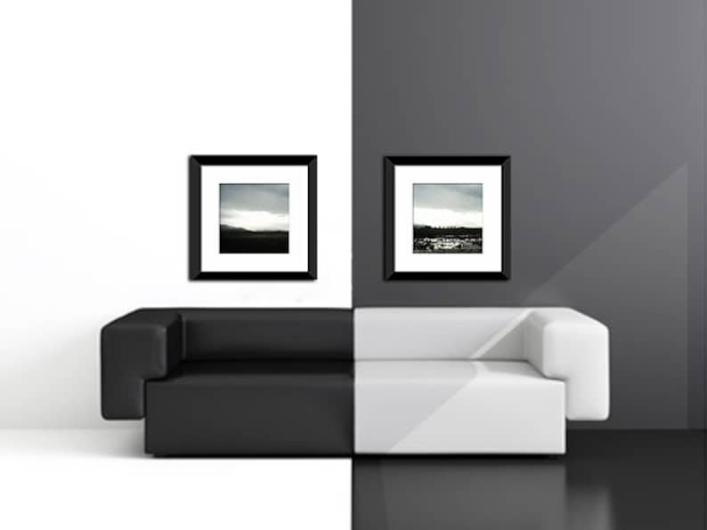 May include: A black leather sofa with a white cushion sits in front of a white wall and a black wall. Two black framed photographs hang on the walls above the sofa.