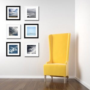 May include: Six black framed photographs of clouds and a sky with birds hanging on a white wall next to a yellow chair.