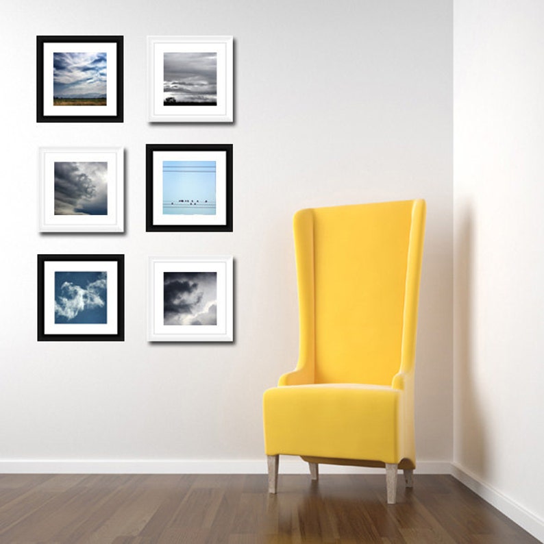 May include: Six black framed photographs of clouds and a sky with birds hanging on a white wall next to a yellow chair.