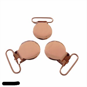 Op de afbeelding: Drie roségoudkleurige metalen clips. Elke clip heeft een cirkelvormig, gewelfd lichaam en een rechthoekige lus. Eén clip is boven de andere twee geplaatst, die in een driehoek zijn gerangschikt.