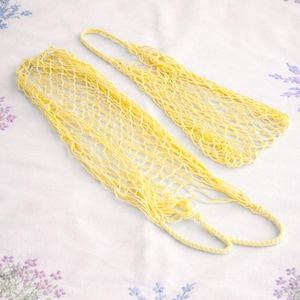 May include: Two yellow, open-weave net bags are displayed on a white floral patterned surface. The bags have long, rectangular bodies with a handle at the top. The bags are made of a soft, woven material.