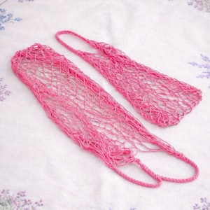 May include: Two bright pink, open-weave net bags with handles, lying on a white surface. The bags are made of a thick, woven material, and the handles are looped. The bags are empty and ready to be filled.