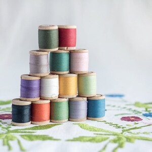 May include: A stack of colorful spools of thread arranged in a pyramid shape on a white fabric with floral embroidery.