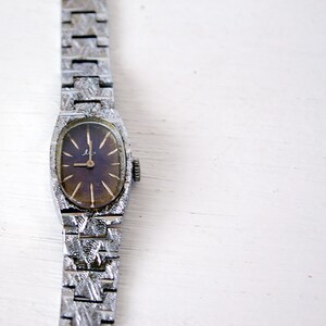 May include: A silver wristwatch with a dark brown face and a patterned metal band.
