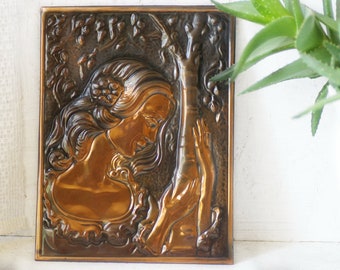 May include: A copper wall plaque with a raised relief design of a woman with long hair leaning against a tree. The plaque has a dark brown patina and is framed in a simple copper frame.