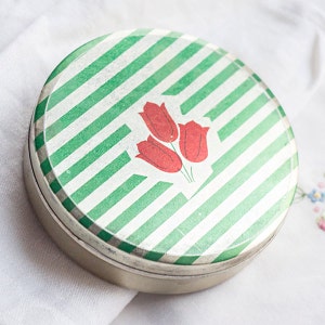 May include: A circular tin with a green and white striped design. The lid has a graphic of three red tulips. The tin is made of metal and looks like a vintage item, possibly antique.