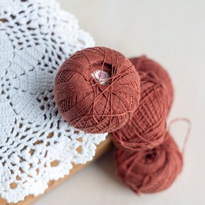 May include: Three balls of rust-colored yarn, one with a white label, on a white crocheted doily.
