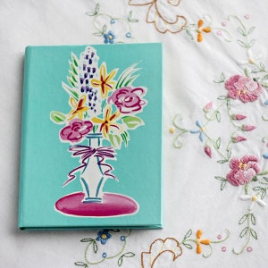 May include: A teal blue notebook with a colorful floral illustration of a bouquet of flowers in a white vase with a pink base. The flowers are yellow, purple, and pink.