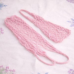 May include: Two pink, open-weave net bags with handles, lying on a white floral patterned surface. The bags are made of a soft, woven material, and the handles are looped. The bags are a light, pastel pink color.