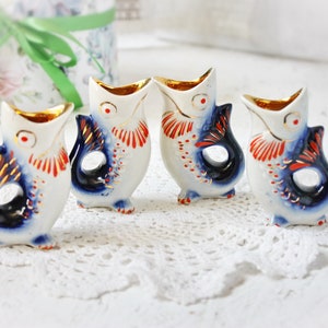 Puede incluir: Cuatro pequeños vasos de chupito de cerámica con forma de pez con detalles pintados en azul, blanco, rojo y dorado. Los vasos están dispuestos en fila sobre una servilleta de encaje blanca.