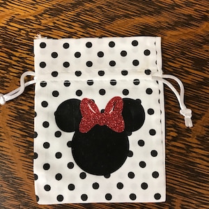 Puede incluir: Una bolsa de cord&oacute;n blanca con lunares negros y una silueta de Minnie Mouse negra y roja.