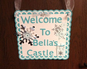 May include: A teal blue welcome sign with a scalloped border. The sign says "Welcome to Bella's Castle" with silver snowflake embellishments and a small white castle cutout.