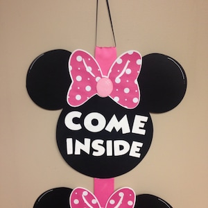 May include: A pink and black Minnie Mouse themed door hanger with the words "Come Inside" in white letters.