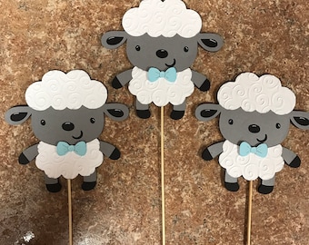 May include: Three gray and white paper sheep with blue bow ties on sticks. The sheep have a white curly design on their bodies.