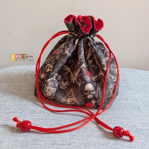 Pode incluir: Uma bolsa de cordão preto com forro vermelho e estampa de caveira e ossos cruzados. A bolsa tem um cordão vermelho com contas vermelhas nas pontas. A bolsa é feita de tecido e tem uma variedade de símbolos e desenhos impressos, incluindo um olho, uma caveira e uma garrafa.