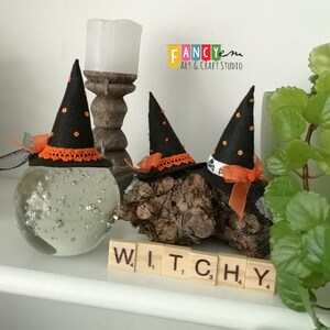 May include: Three black witch hats with orange and white trim, sitting on a wooden log and a glass orb. The hats are decorated with black polka dots and a black and white ribbon. The word "WITCHY" is spelled out in wooden letter tiles.