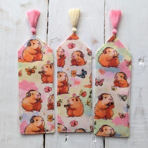 May include: Three decorative bookmarks featuring a guinea pig design. Each bookmark shows a guinea pig wearing headphones and holding an object, set against a background of butterflies and flowers. The bookmarks have pink, cream, and peach tassels.